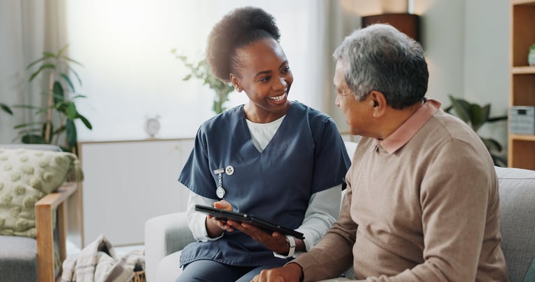 Young African American female caregiver with tablet providing telehealth support to senior man on sofa at home