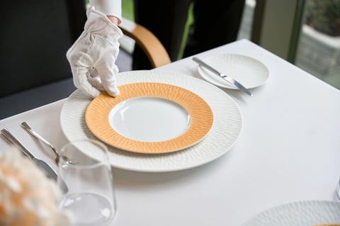 Fine dining table setting with white plate rimmed in orange, folded napkin, silverware, and glass on white tablecloth
