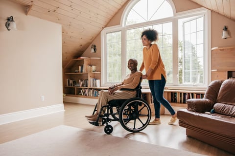Caregiver assisting elderly woman in wheelchair in bright, modern living room with wooden beams and large windows